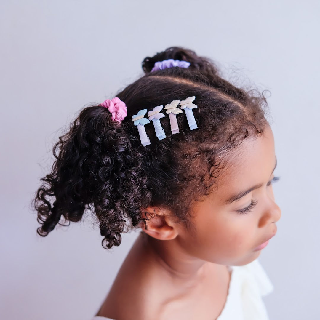 Zijaanzicht van een kind met donker krullend haar, versierd met drie mimi & lula vlinder haarclipjes en een roze scrunchie.