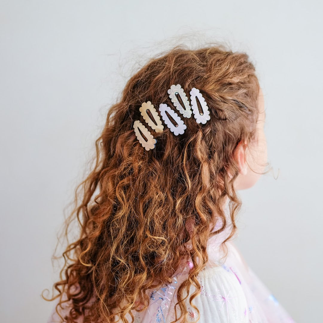 Een kind met lang, krullend bruin haar draagt drie mimi & lula mini scalloped haarclipjes aan de zijkant.