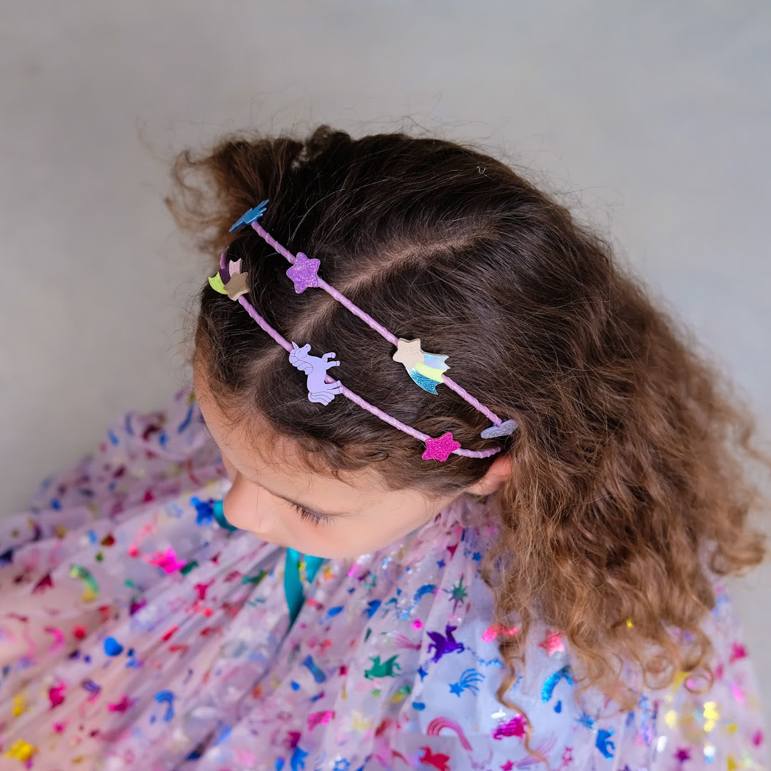 Een kind draagt de roze kinder hoofdband met eenhoorn en ster versieringen in bruin krullend haar.