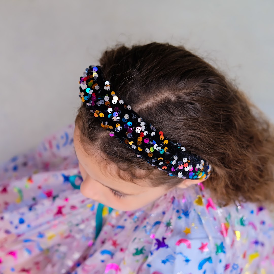 Bovenaanzicht van een kind met de mimi & lula hoofdband met veelkleurige lovertjes in bruin krullend haar.
