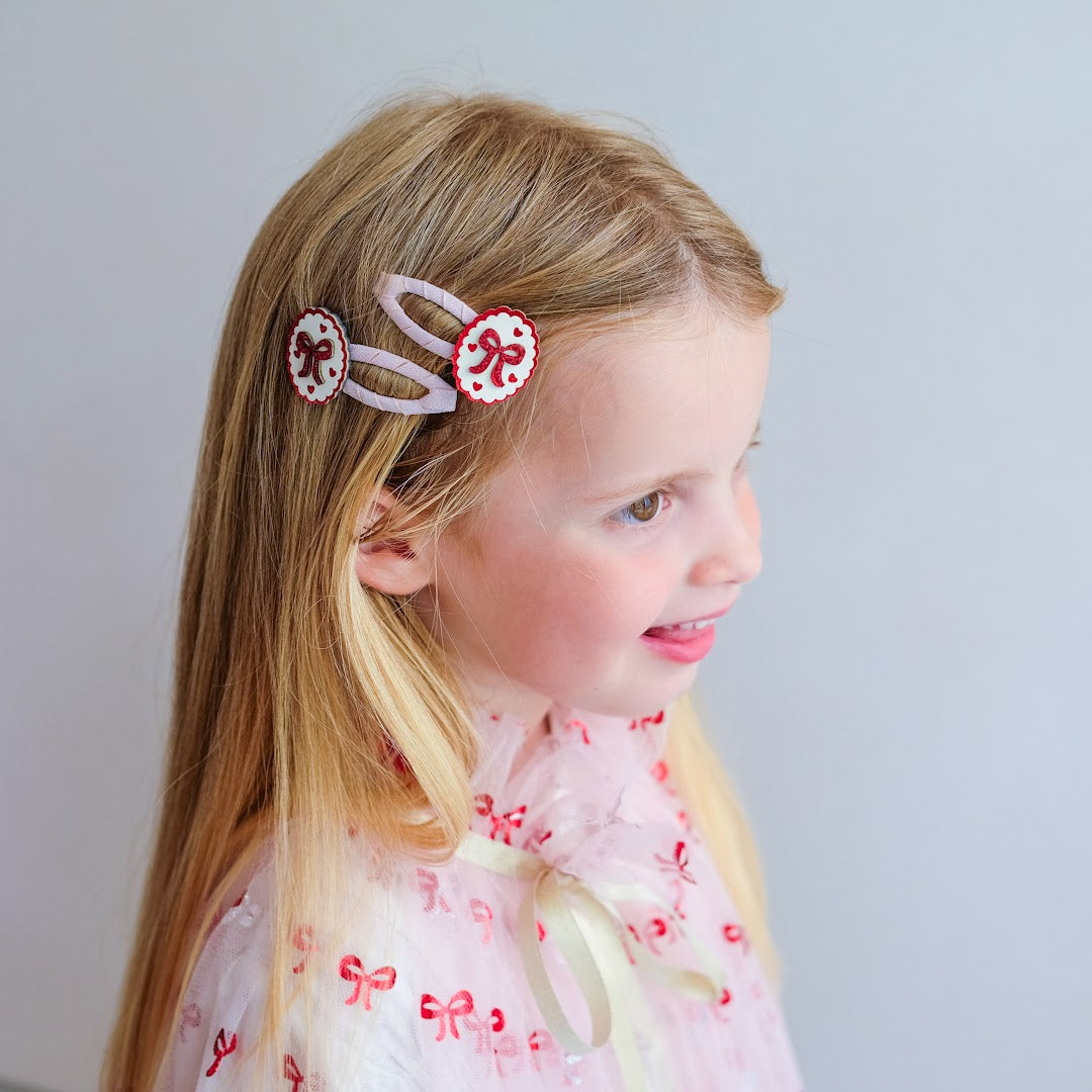 Een lachend meisje met blond haar draagt twee mimi & lula haarclipjes met strikjes en hartjes.