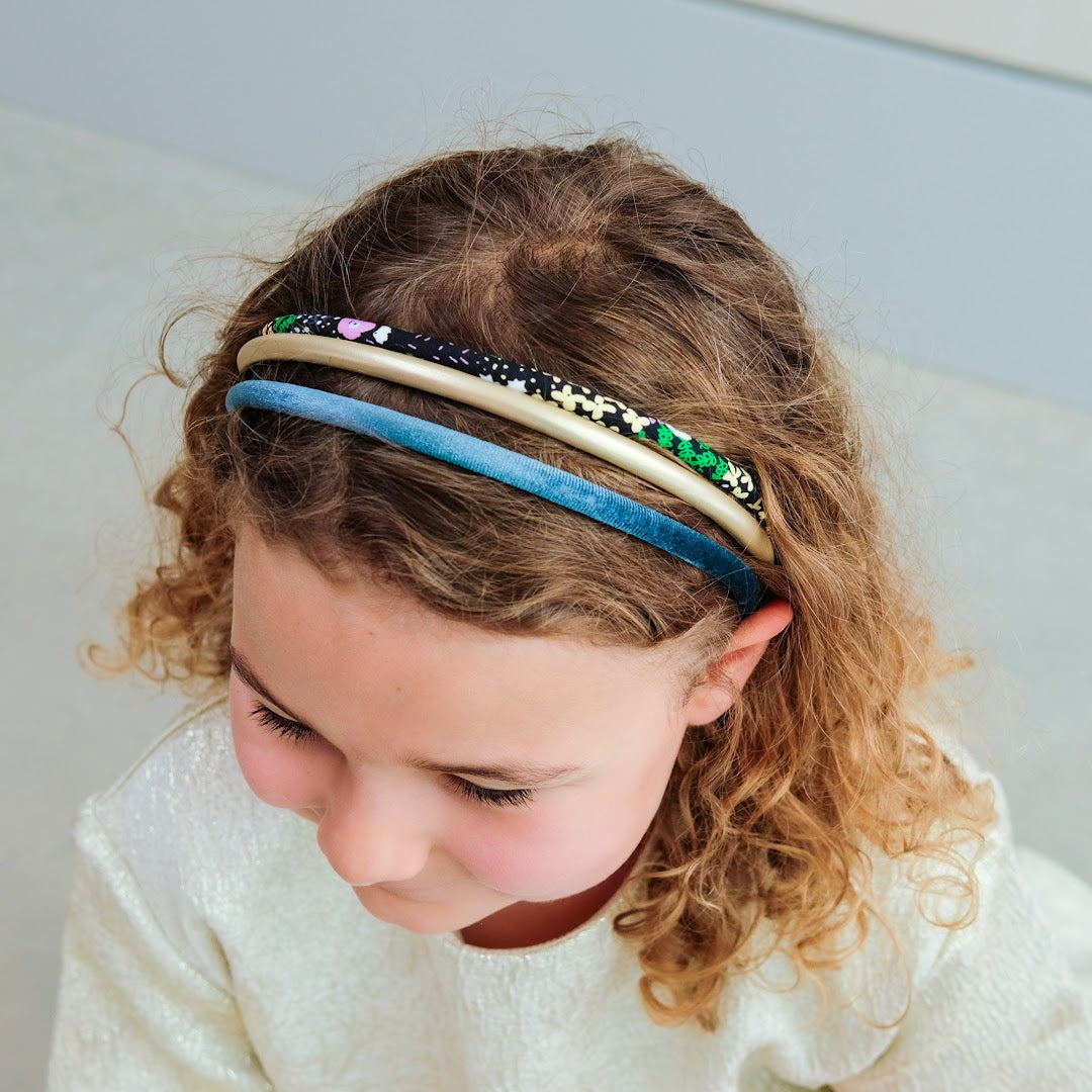 Een kind met krullend haar draagt drie mimi & lula kinderhaarbanden: een goudkleurige, een teal fluwelen en een met bloemenpatroon.