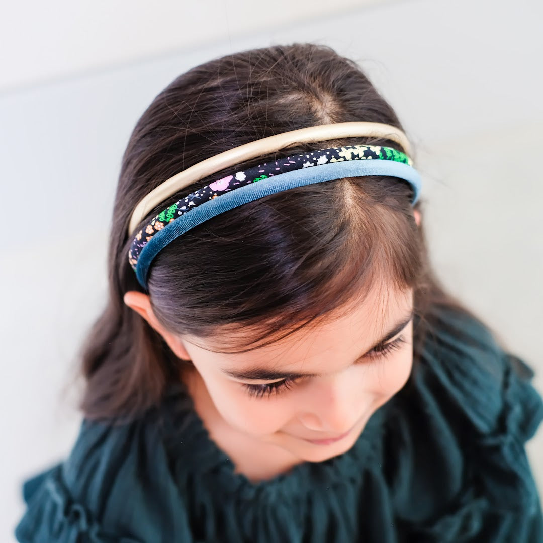 Een kind met donker steil haar draagt drie mimi & lula kinderhaarbanden: een goudkleurige, een teal fluwelen en een met bloemenpatroon.