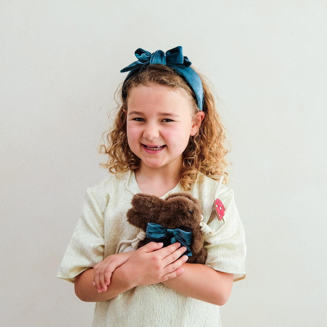 Lachend meisje met krullen draagt de blauwe fluwelen hoofdband met strik en houdt een knuffel vast.