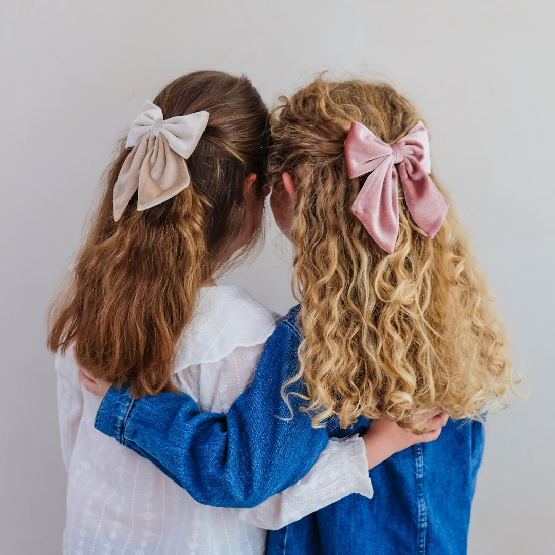 Twee meisjes met lange haren dragen de mimi & lula Agnes haarstrikken, één beige en één roze.