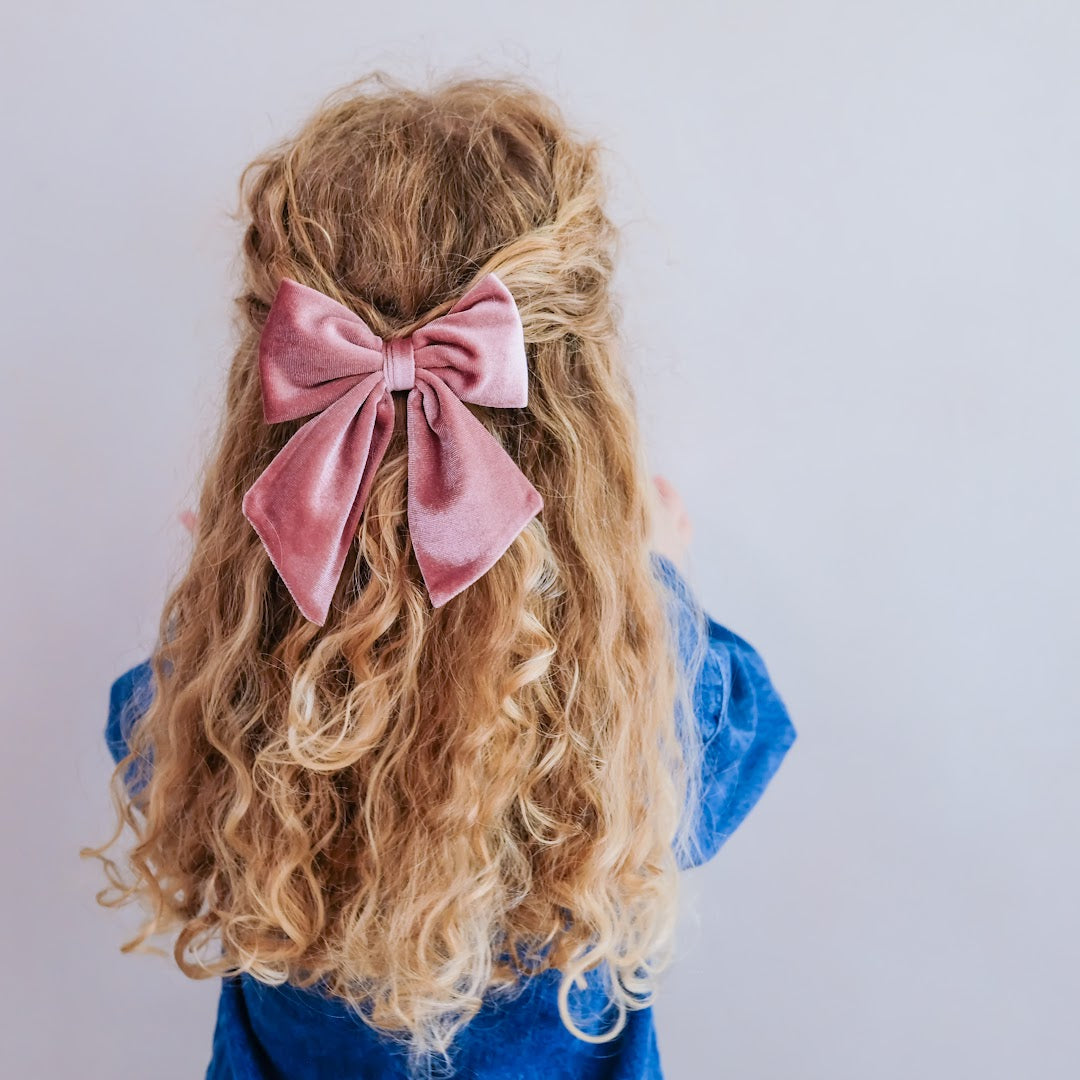 Een meisje met lang blond krullend haar draagt een grote roze mimi & lula Agnes fluwelen haarstrik.