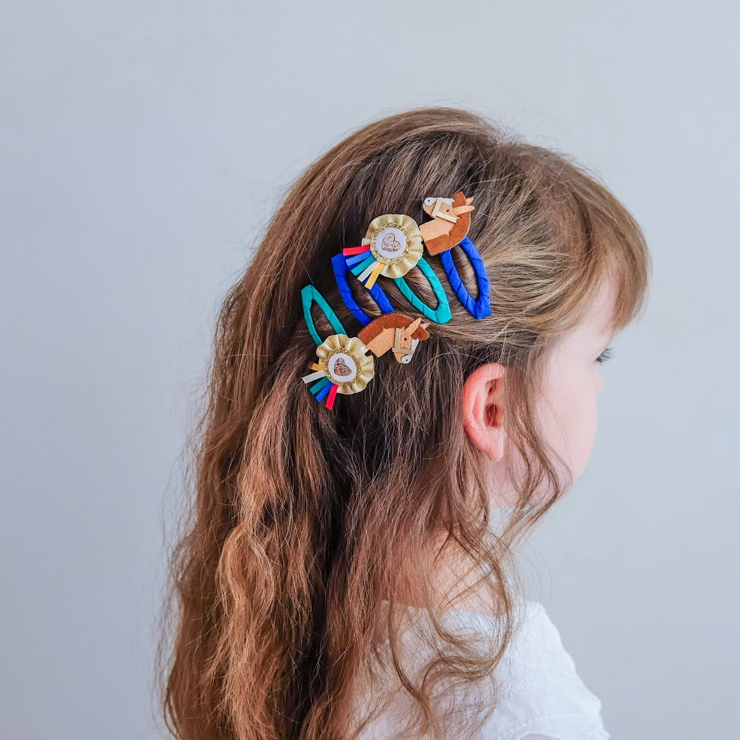 Een kind met lang bruin haar draagt mimi & lula haarclipjes met paarden en rozetten.