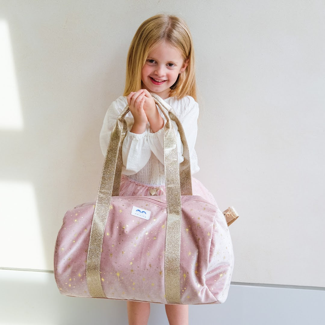 Een lachend meisje houdt de mimi & lula roze fluwelen weekendtas met gouden sterren voor zich.