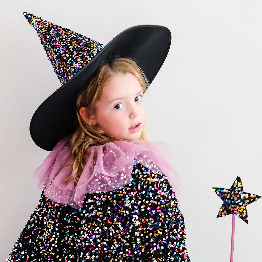 Kind draagt een mimi & lula heksenhoed en cape met veelkleurige pailletten, met een toverstaf.