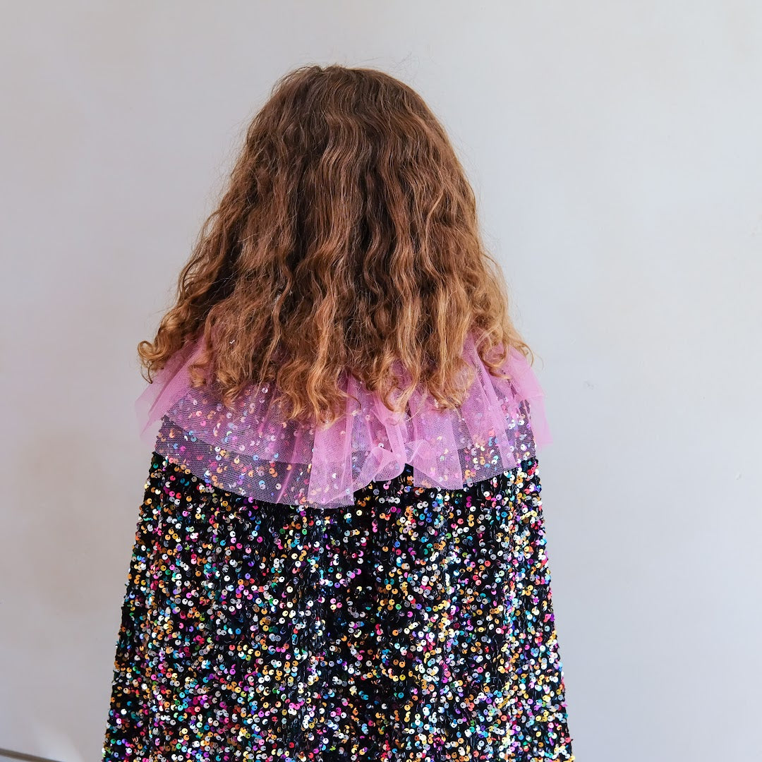 Kind van achteren gezien met lange krullen, draagt de verkleedcape met kleurrijke pailletten en roze tule kraag.