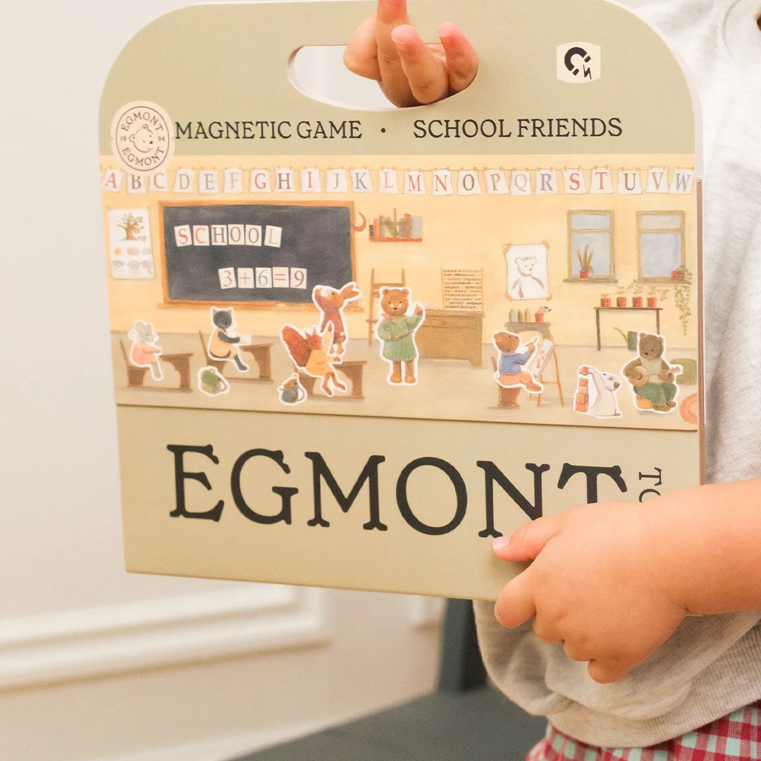 Kinderhanden houden de verpakking van het Egmont Toys magnetisch spel School Friends vast, met een schoolscène en dierenfiguren.