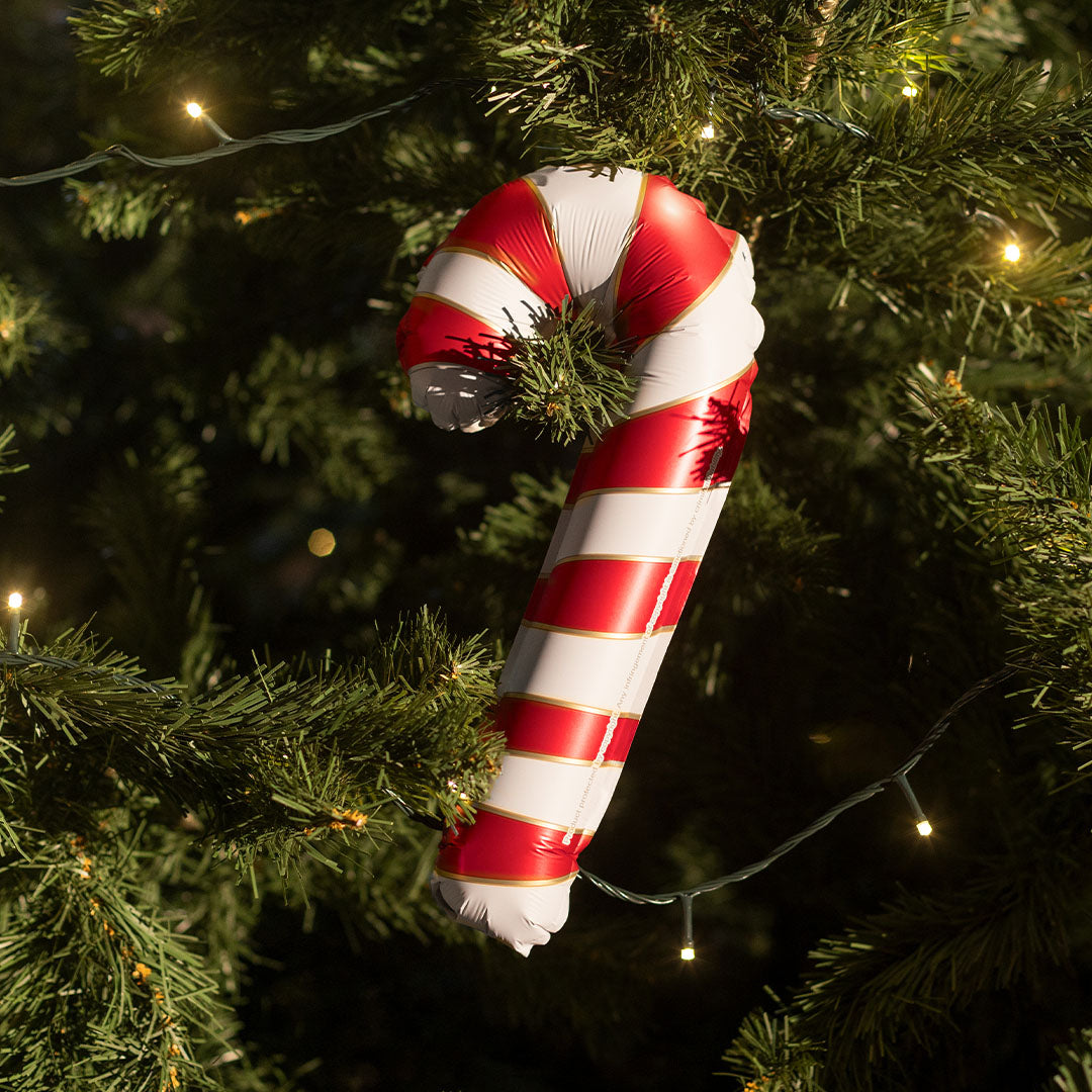 Partydeco folieballon in de vorm van een rood-wit gestreepte zuurstok hangend in een kerstboom met lichtjes.