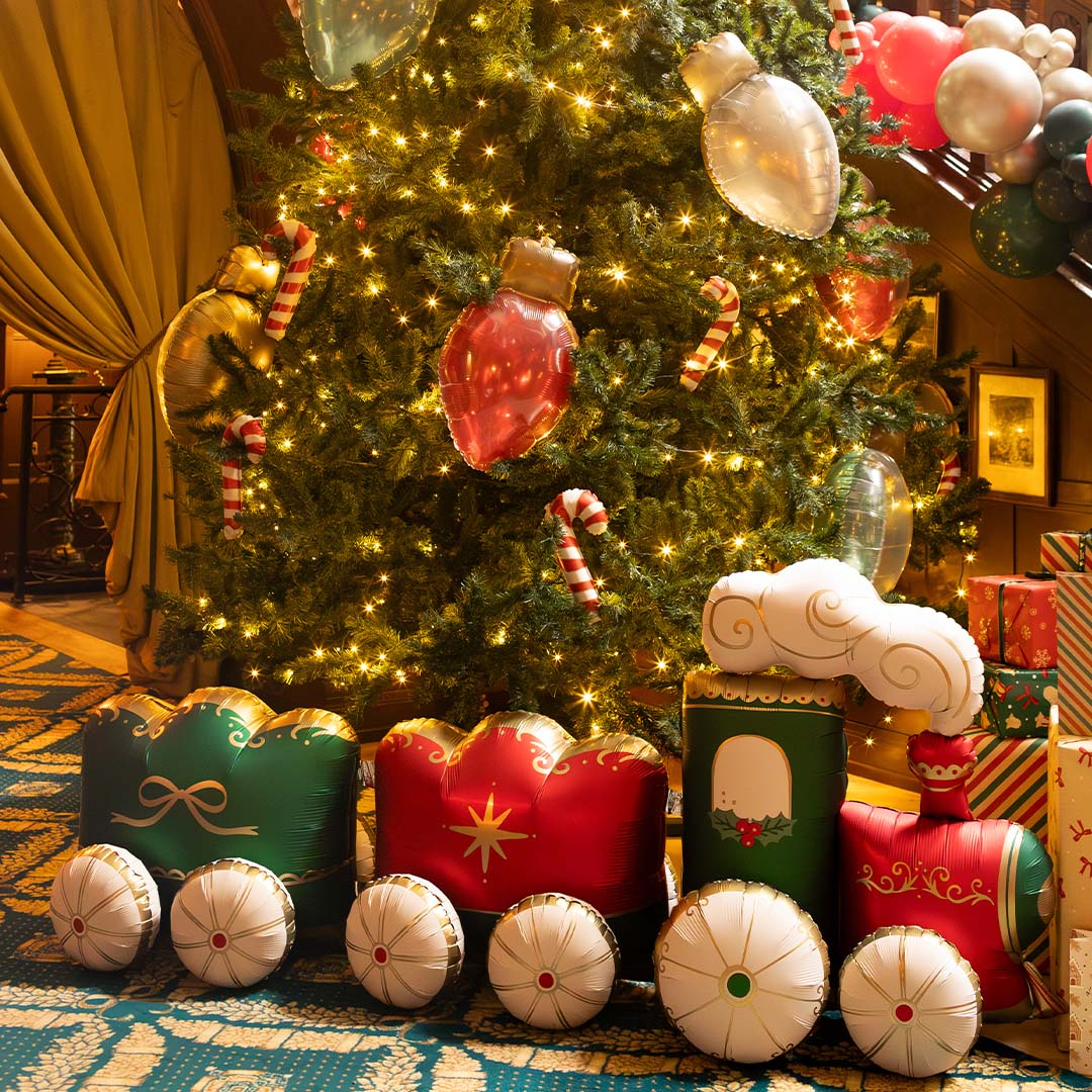 De partydeco XXL folieballon kersttrein staat als feestelijke decoratie voor een verlichte kerstboom.