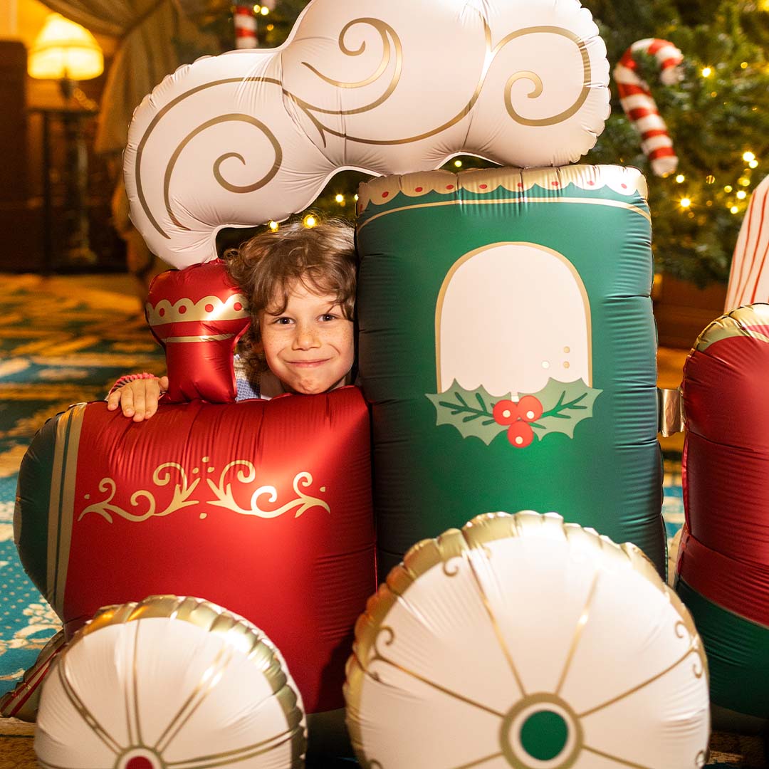 Een lachende jongen kijkt over de partydeco XXL folieballon kersttrein heen, die voor een kerstboom staat.