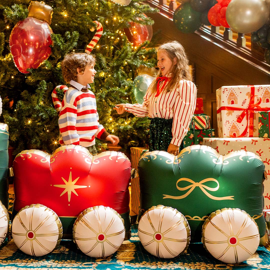 Twee lachende kinderen staan naast de partydeco XXL folieballon kersttrein voor een kerstboom met cadeaus.