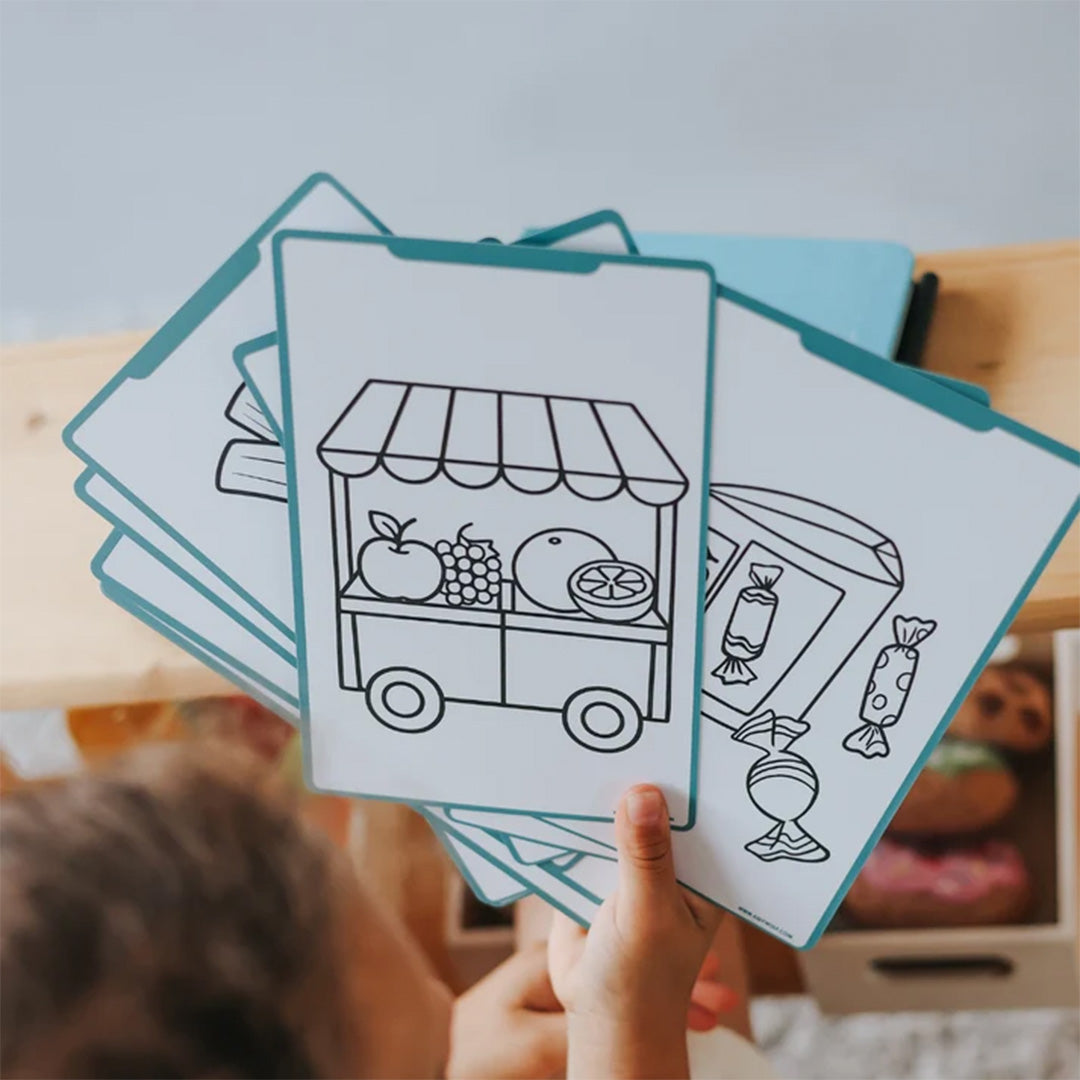 Een kinderhand houdt meerdere witte tekensjablonen vast met lijntekeningen van een marktkraam en snoepjes.
