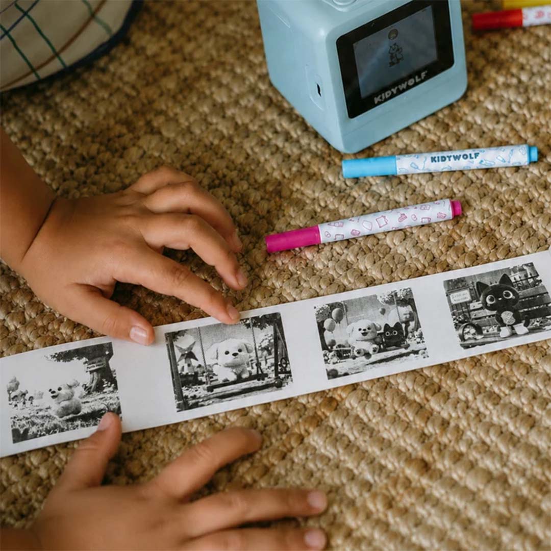 Kinderhanden houden een afgedrukte strook met zwart-wit dierenfoto's vast, naast de Kidywolf Kidyprinter AI en twee stiften.