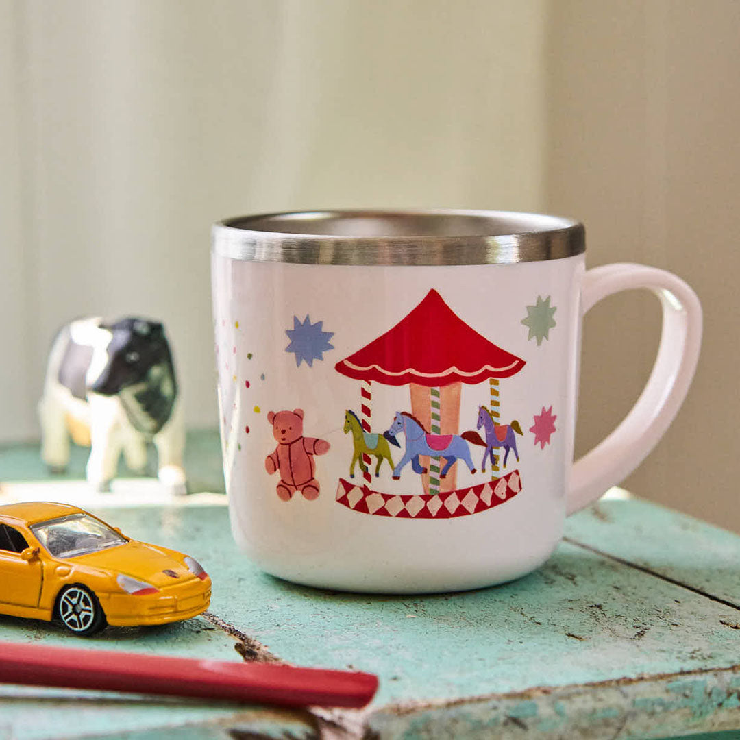 rice roze kinderbeker met een carrousel en teddybeer, staand op een blauwe tafel naast speelgoed.