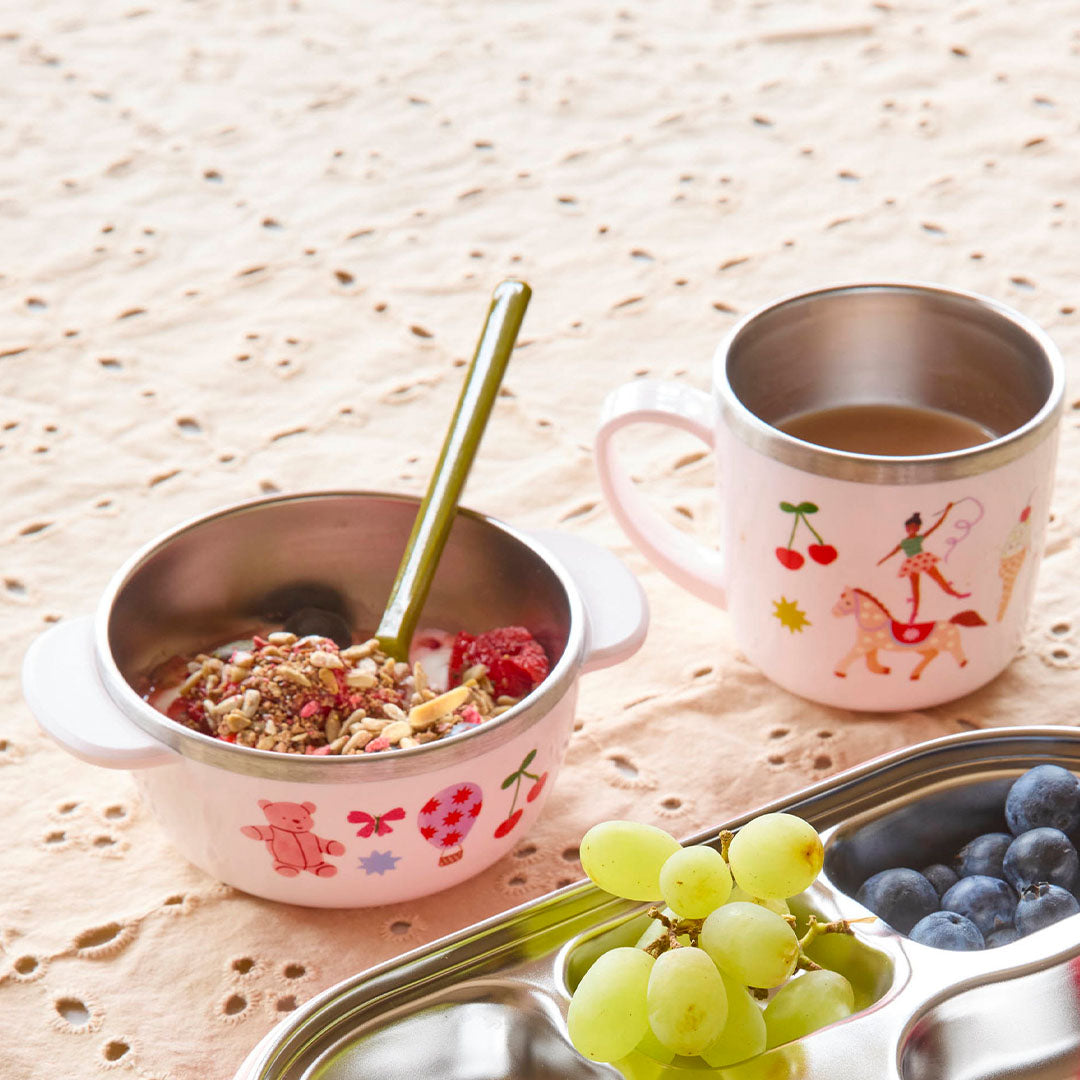 rice roze rvs kinderkom met ontbijt, beker en fruit op een tafelkleed.
