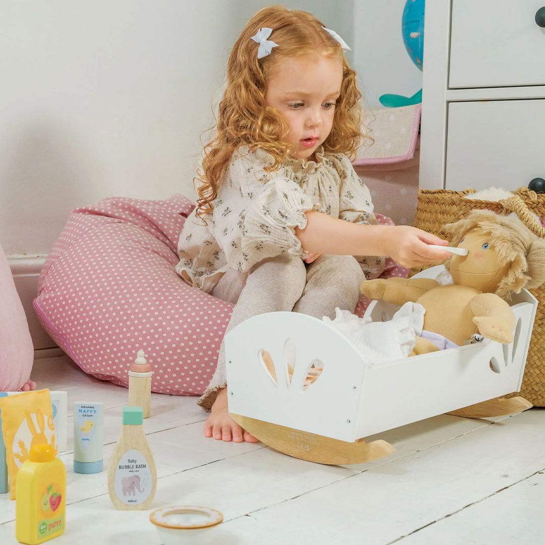 Een meisje voedt een pop in een wit wiegje met een lepel uit de Tender Leaf Toys poppenverzorgingsset.