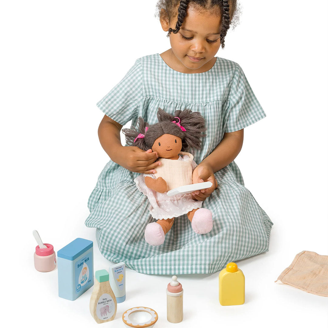 Een meisje zit op de grond en houdt een pop vast met een lepel uit de Tender Leaf Toys poppenverzorgingsset.
