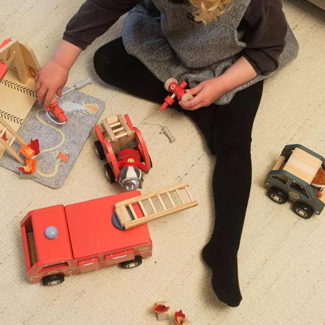 Een kind speelt op een vloerkleed met de Egmont Toys houten brandweerwagen speelset en een knuffeldas.