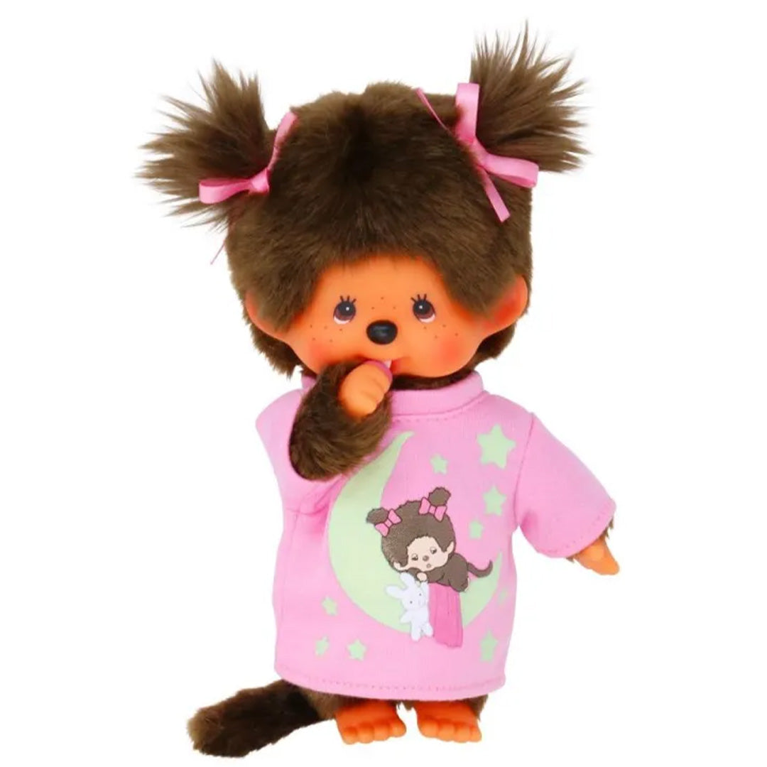 Monchhichi meisje pop in roze pyjama met maan- en sterrenpatroon, met handje bij de mond.