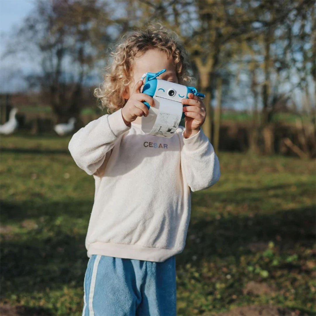 Een kind met krullen houdt de blauw-witte kidysnap kindercamera voor het gezicht in een groene omgeving.