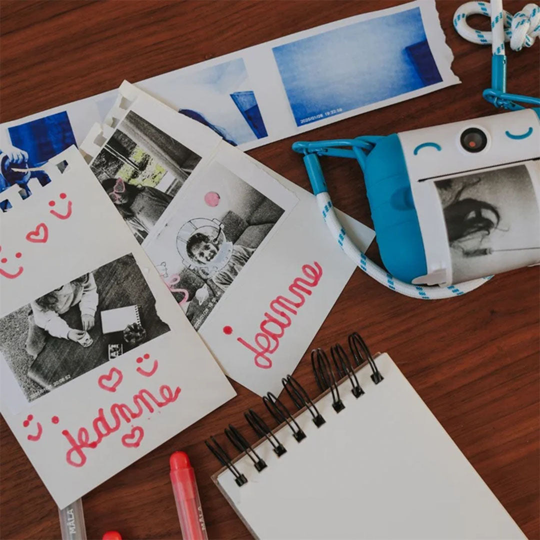 De blauw-witte kidysnap kindercamera ligt op een tafel met geprinte foto's en een notitieboekje.
