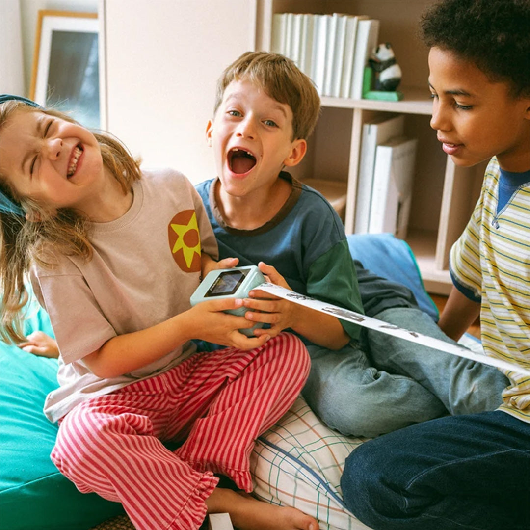 Drie lachende kinderen spelen samen met de Kidywolf Kidyprinter AI speelgoedprinter die een lange strook papier afdrukt.