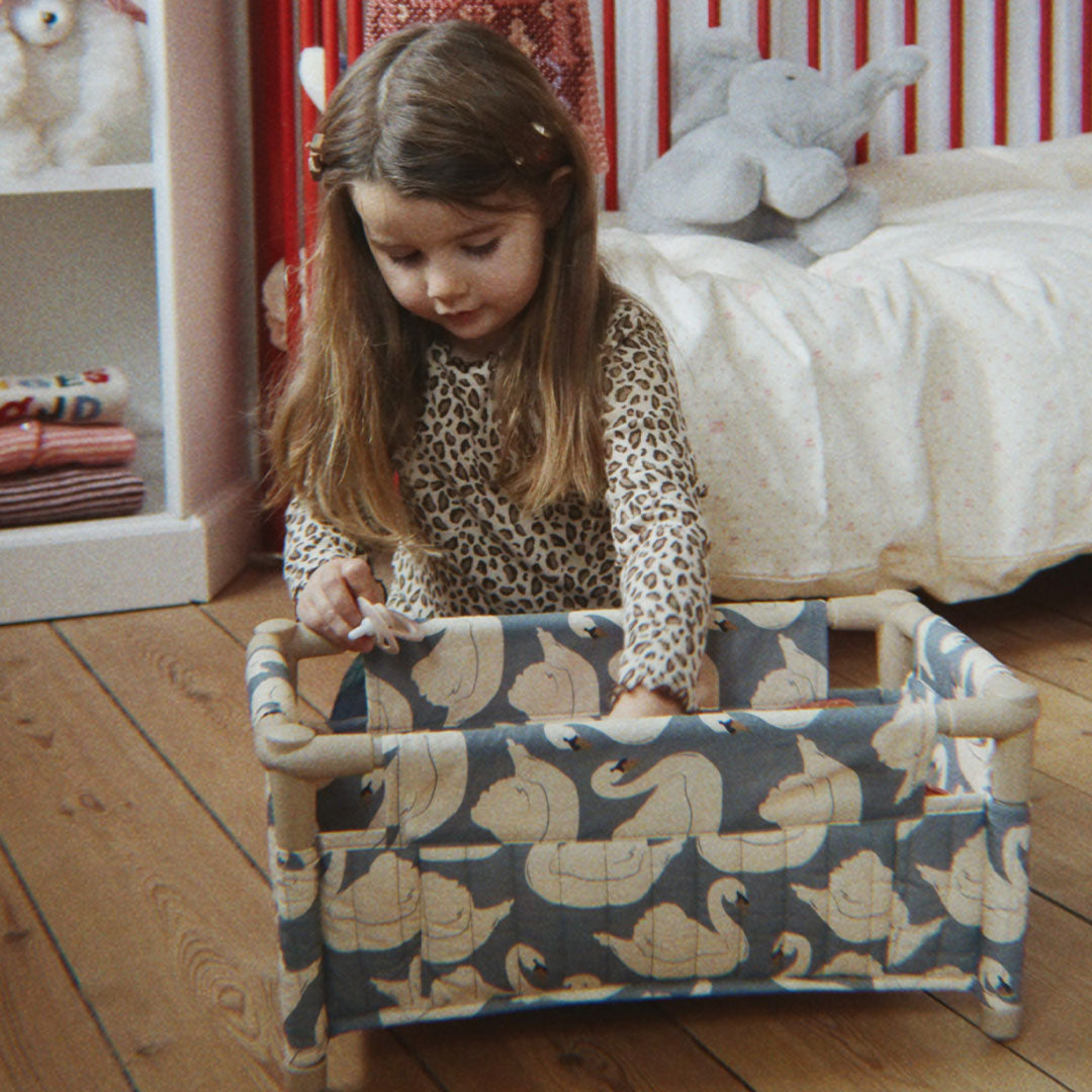 Een meisje speelt met het Konges Sløjd poppenbed met blauwe stof en zwanenpatroon in een kinderkamer.