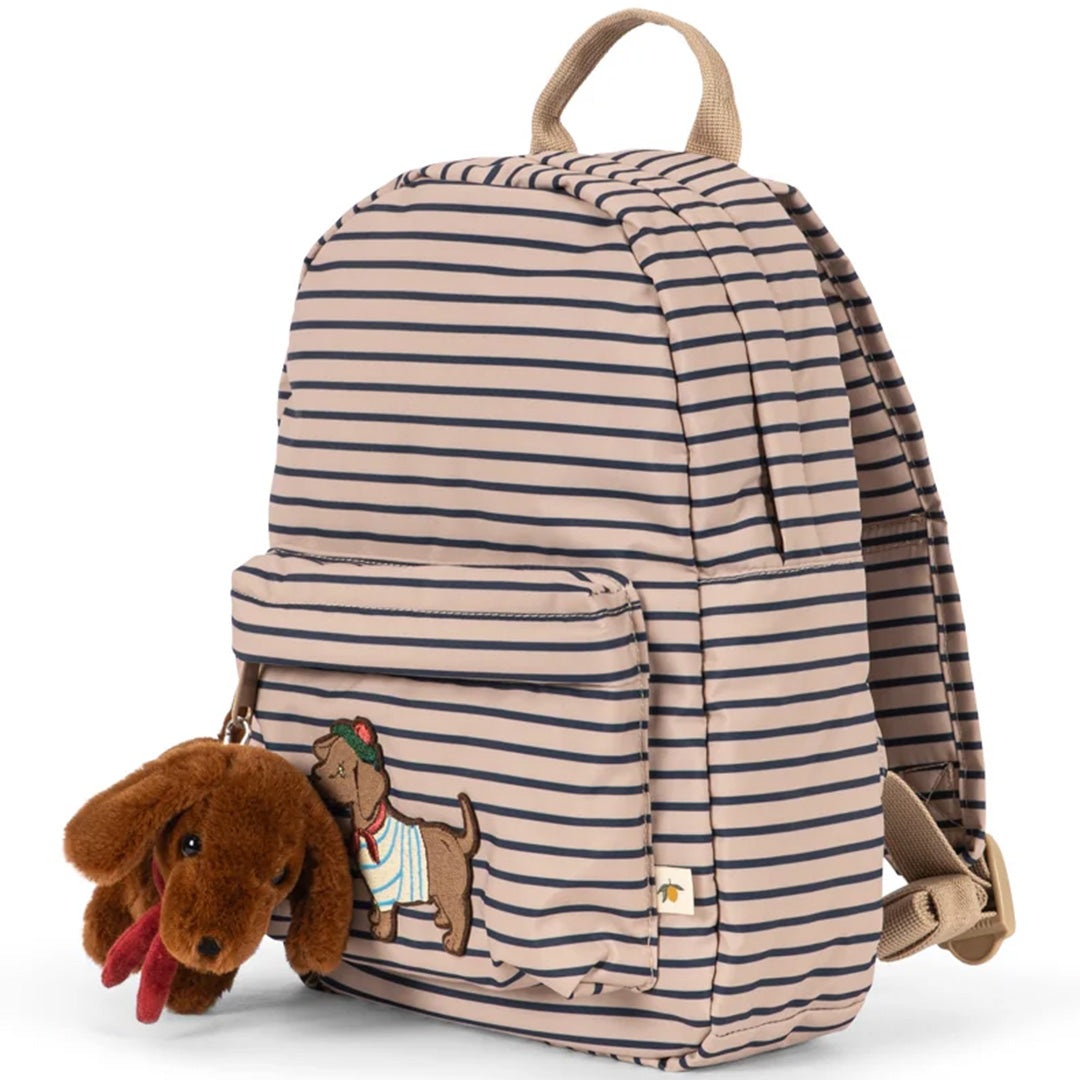Konges Sløjd peuterrugzak met beige en marineblauwe strepen, een geborduurde hond en een pluche hond sleutelhanger, schuin van voren gefotografeerd.