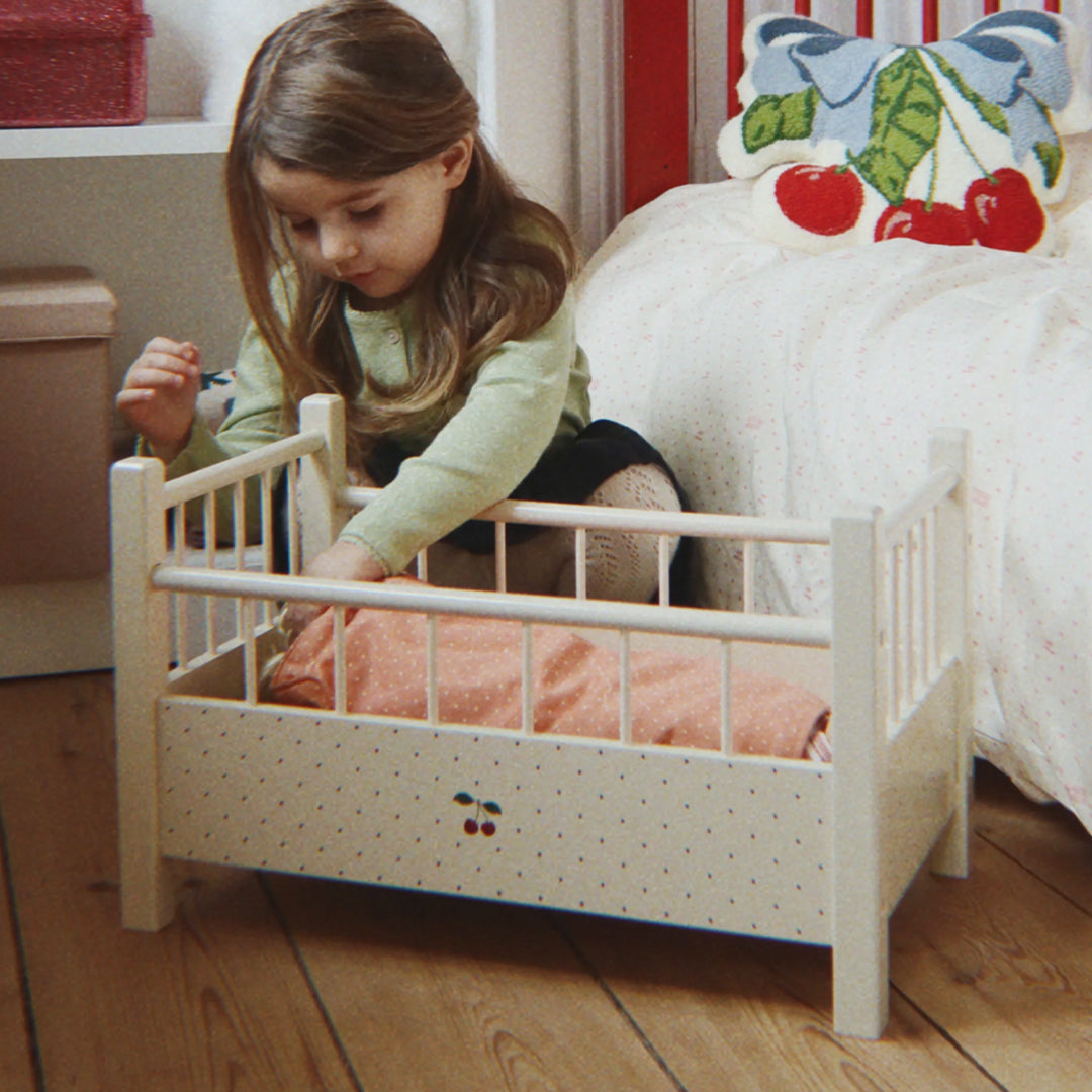 Een meisje speelt met het Konges Sløjd houten poppenbed, dat beige is met stippen en een kersenillustratie.