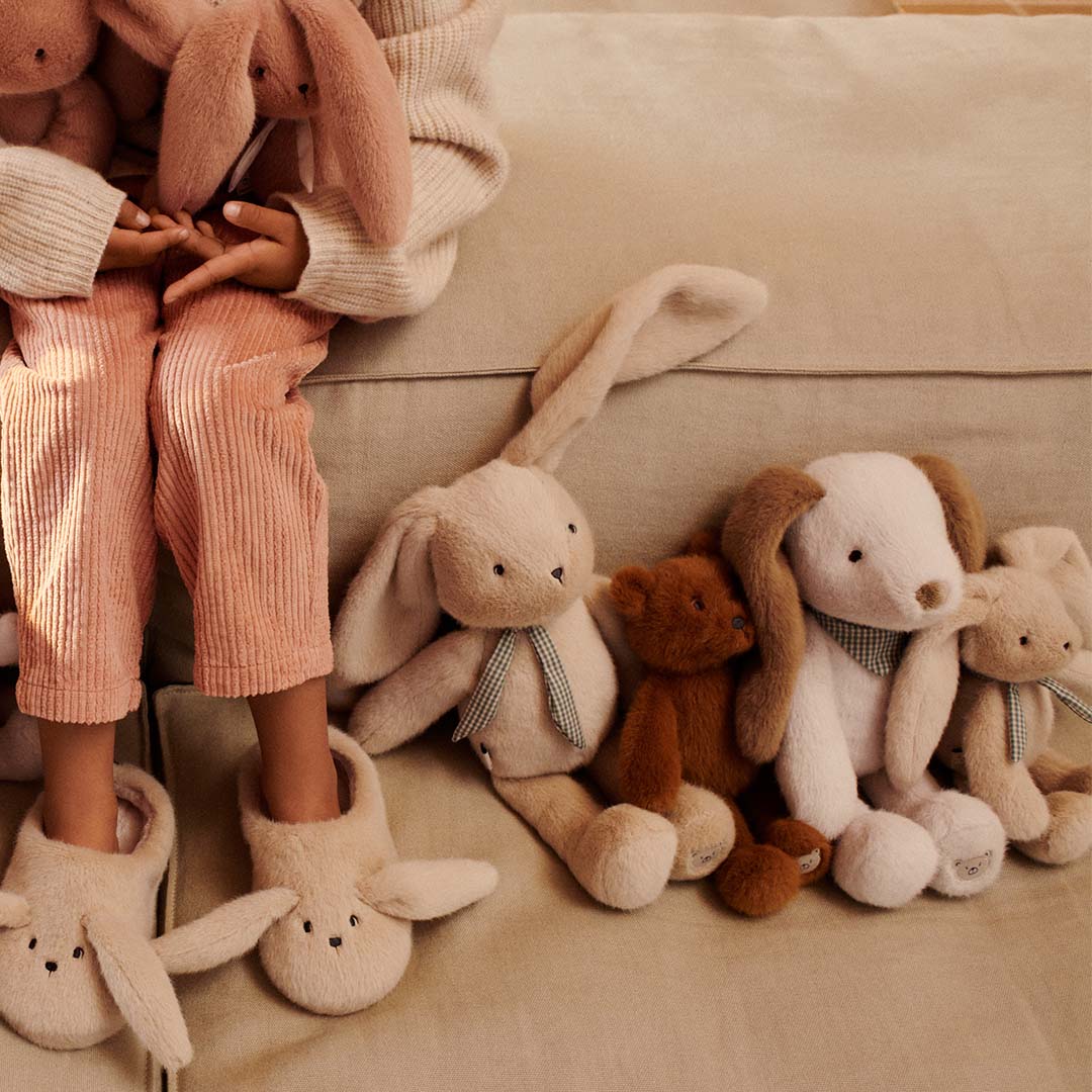 Een kind zit op een bank tussen een collectie pluchen knuffels, waaronder konijnen en een beer.