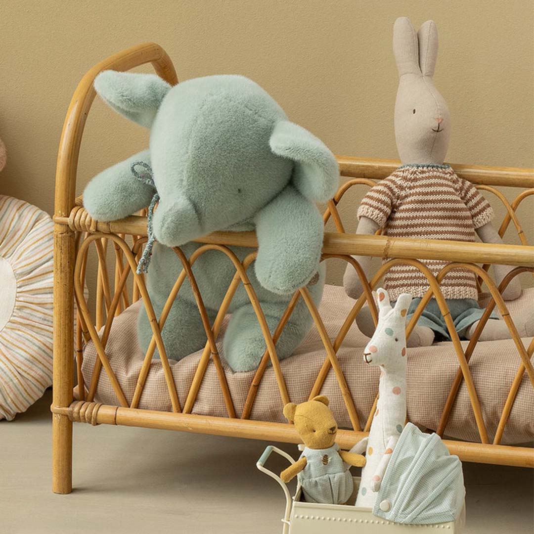 De Maileg pluchen knuffelolifant hangt over een rieten poppenbedje met andere knuffels.