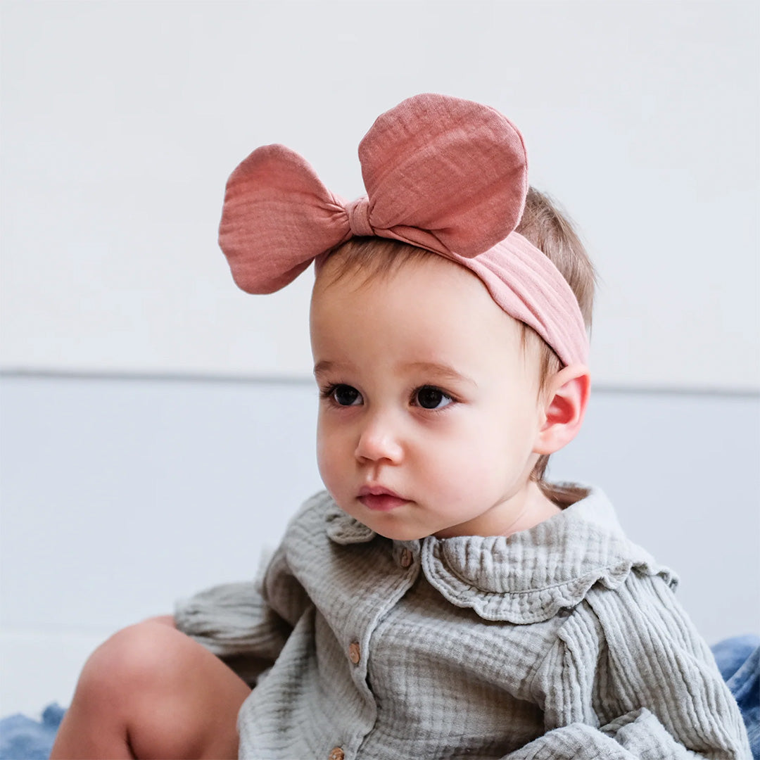 Een baby met een grijze blouse draagt de roze mimi & lula babyhaarband met grote strik.