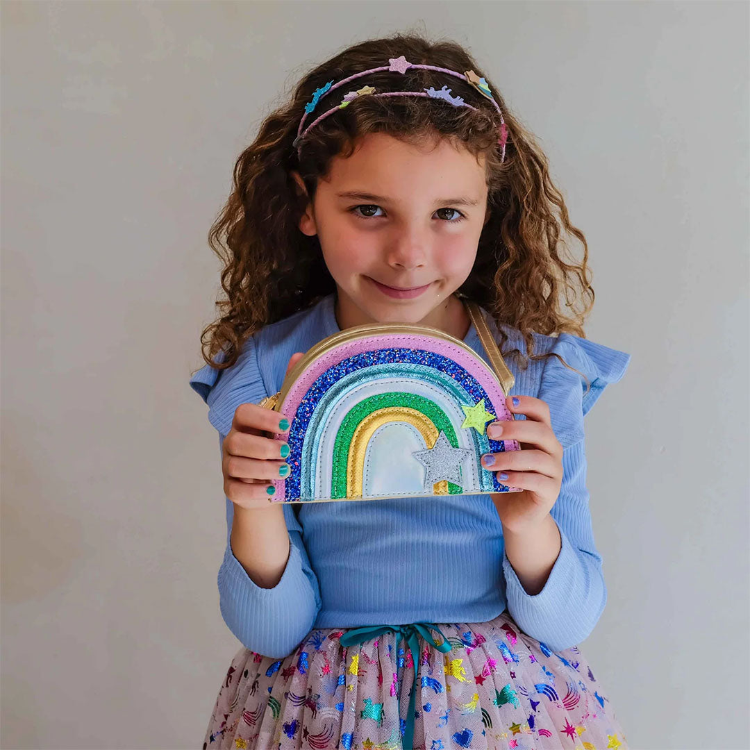 Een lachend meisje met krullend haar toont het mimi & lula regenboog schoudertasje met glitter en sterren.