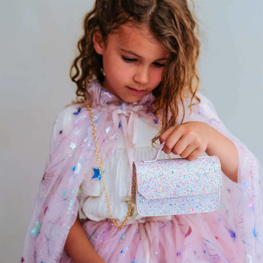 Een meisje draagt het Mimi & Lula roze glitter schoudertasje met een gouden ketting en een glinsterende cape.