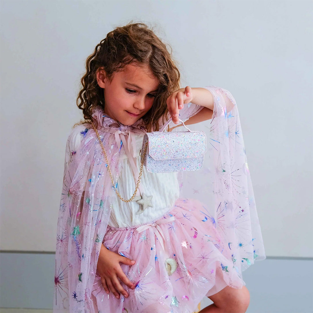 Een meisje draagt de mimi & lula roze verkleedcape met sterren en manen en houdt een glittertasje vast.
