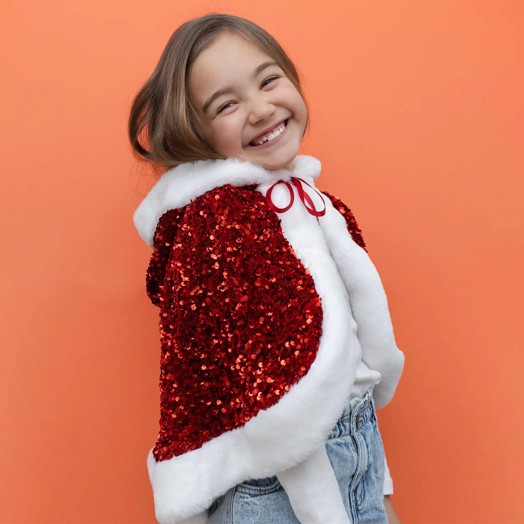 Lachend meisje draagt de mimi & lula rode Mrs Claus verkleedcape met pailletten en witte bontrand, tegen een oranje achtergrond.