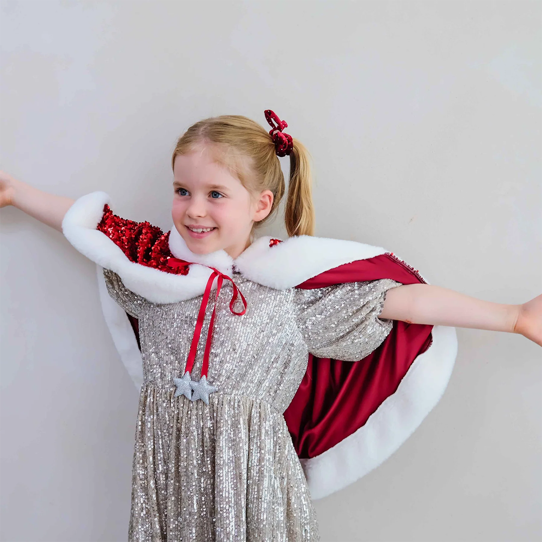 Meisje met blonde staartjes draagt de mimi & lula rode Mrs Claus verkleedcape met pailletten en witte bontrand, met uitgestrekte armen.