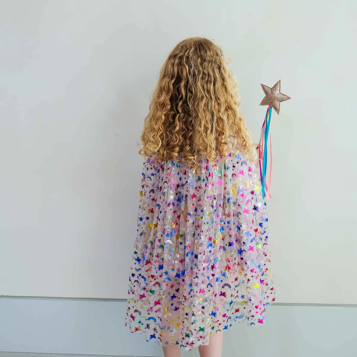 Een meisje van achteren gezien, draagt de Mimi & Lula verkleedcape met regenboog eenhoorn motief en houdt een sterrenstaf vast.