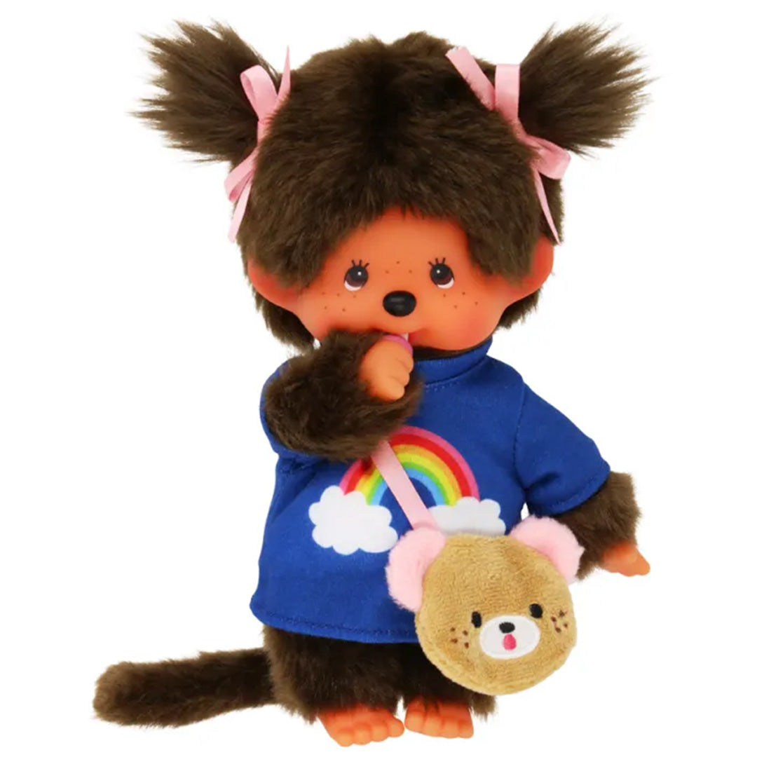 Monchhichi meisje pop met bruine vacht, roze strikjes, een blauw shirt met regenboog en een tasje, met een hand bij de mond.