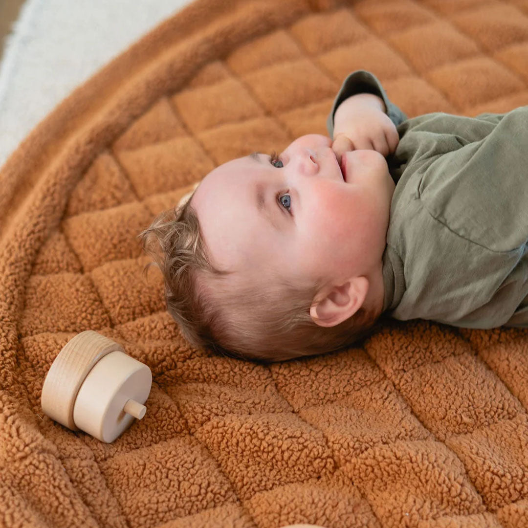 Een baby ligt op het play&go bruine teddy speelkleed en kijkt omhoog, met een houten speeltje ernaast.