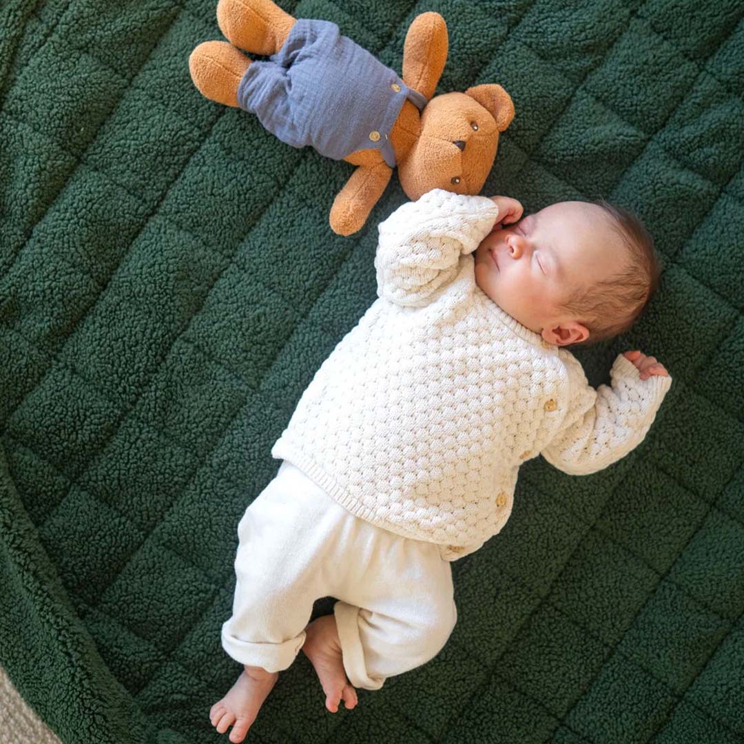 Slapende baby ligt op een donkergroen teddy babyspeelkleed naast een teddybeer.