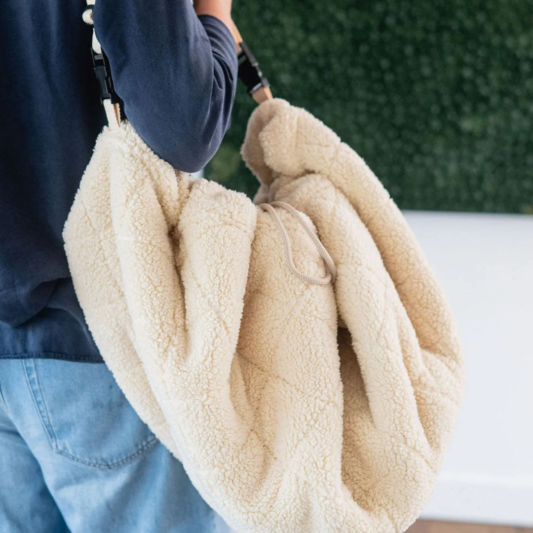 Een persoon draagt de beige play&go babyspeelkleed tas van teddy stof over de schouder.