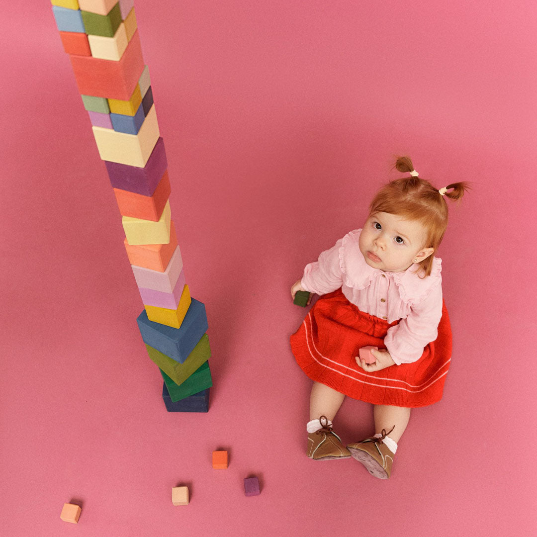 Een baby kijkt omhoog naar een hoge toren van Raduga Grëz veelkleurige houten bouwblokken op een roze achtergrond.