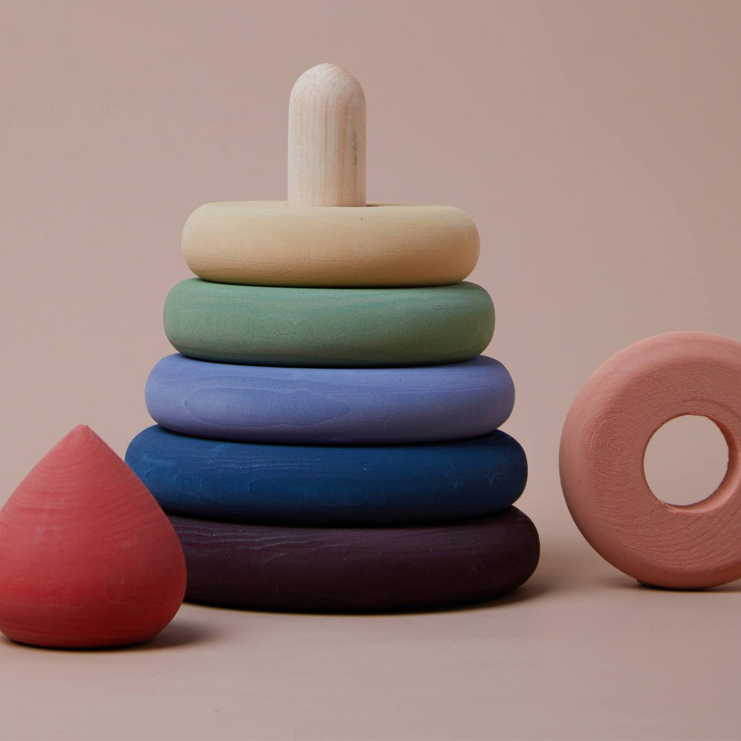 Raduga Grëz houten stapeltoren voor baby's met regenboogkleurige ringen en een natuurhouten pin, deels gedemonteerd op een lichtbruine achtergrond.