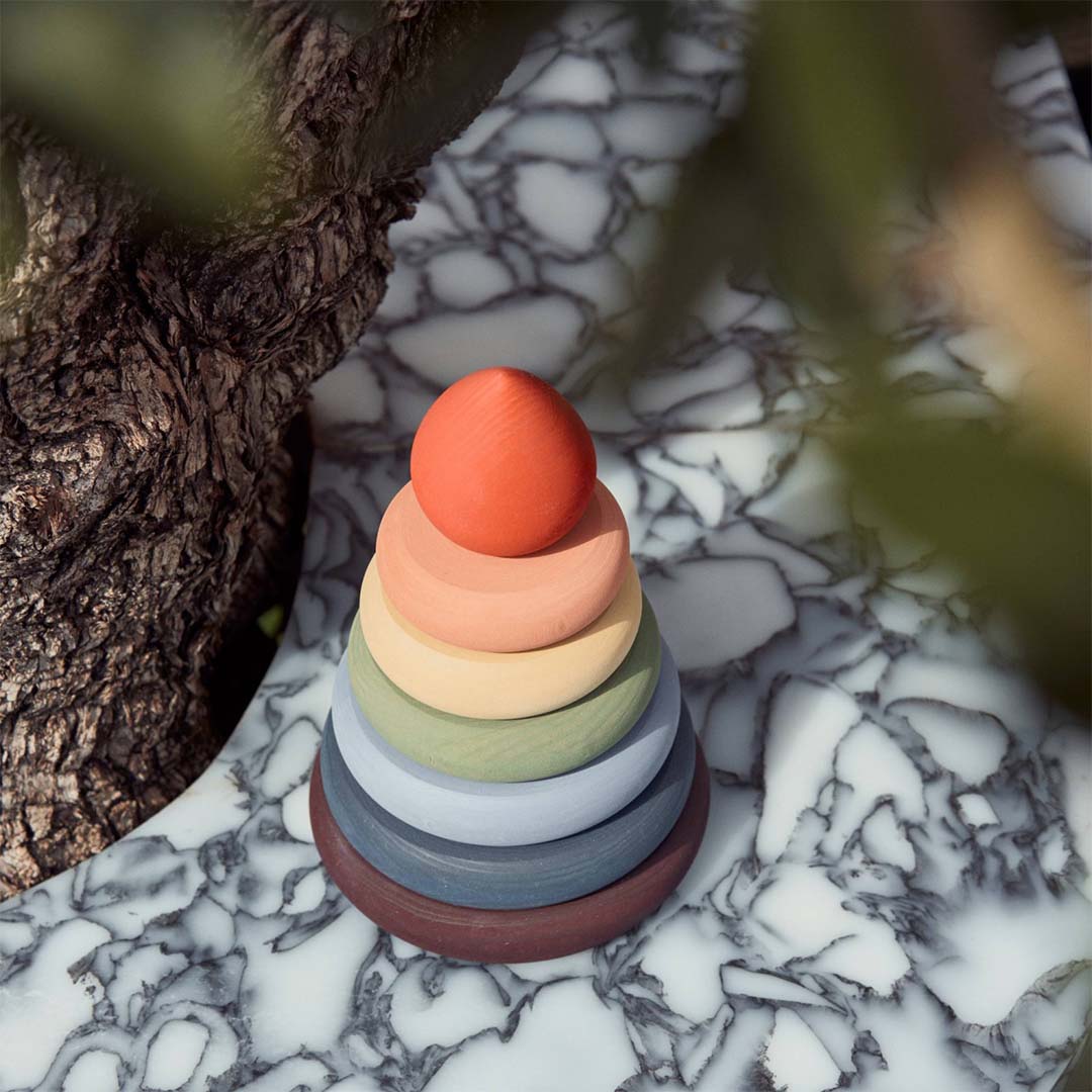 Raduga Grëz houten stapeltoren voor baby's met gedempte regenboogkleurige ringen en een oranje top, op een marmeren ondergrond met een boomstam en bladeren.
