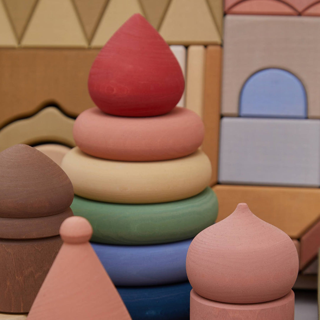 Raduga Grëz houten stapeltoren voor baby's met regenboogkleurige ringen en een rode top, omringd door andere houten speelgoedblokken in vergelijkbare kleuren.